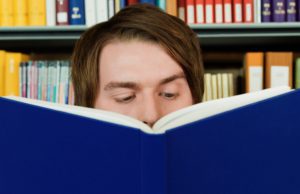 A young man reading a book with his lower half of his face covered with a book.