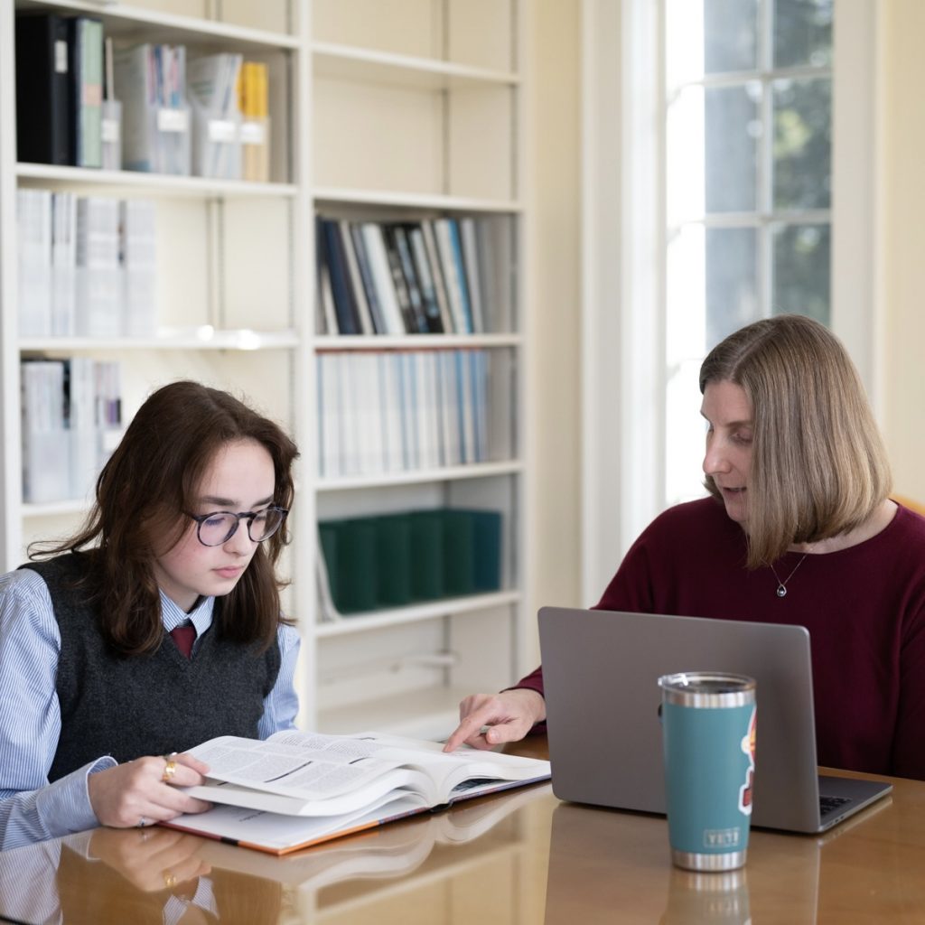 A high school teacher and student are reviewing a textbook side by side while seated in a library.