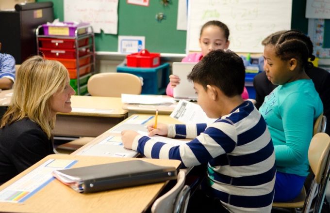 A boy in a striped shirt sits at a table while a teacher helps him