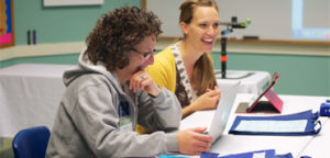 two smiling women at a table looking at their laptops