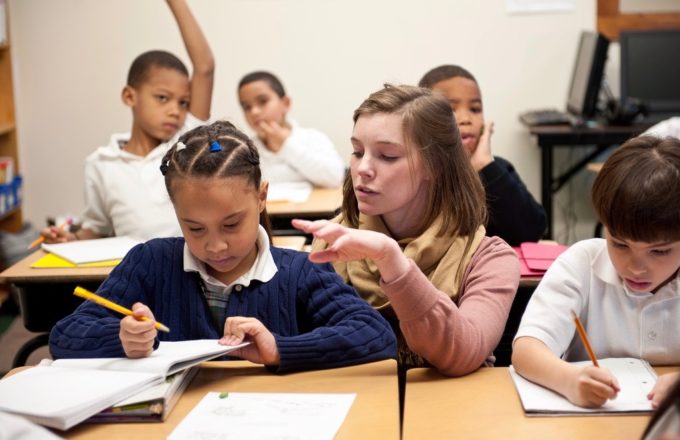 A teacher helping a student while other students in the background have their hands raised