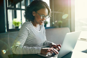 Young African woman wearing glasses sitting by herself at a table using a laptop to browse online