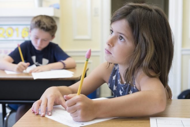 A young girl writing with a pencil at a desk in a classroom.