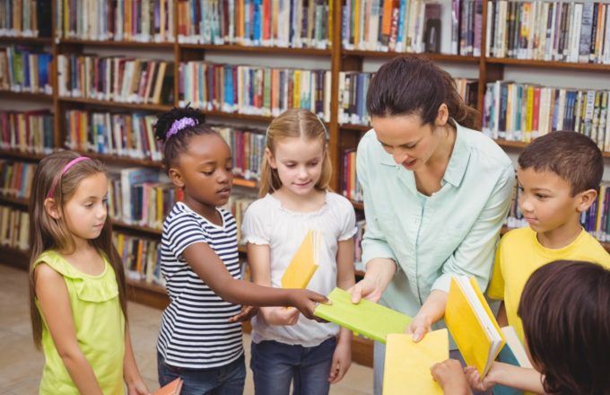 Elementary students are selecting books in a library alongside their teacher.