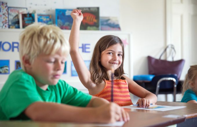 Two students in a classroom with a girl raising her hand