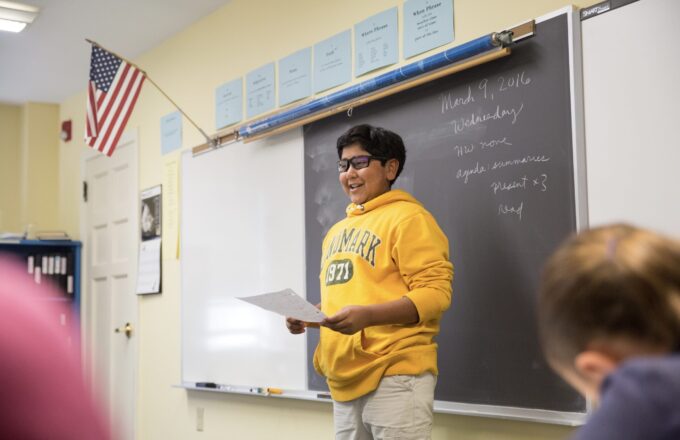 A high school student is presenting to the class at the front of the room.