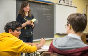 A teacher stands in front of a chalk board and speaks with students
