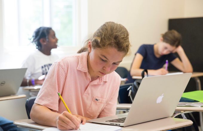 A young girl writes at a desk with an open computer