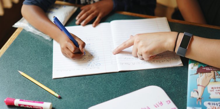 Close-up view of a young student's hands writing in a lined notebook with a blue pen, guided by an adult's pointing finger, which is directing the student on the text. The notebook lays on a green desk, and a pink marker, a yellow pencil, and another notebook with a cartoon cover are also visible, suggesting a lively and engaging learning environment.