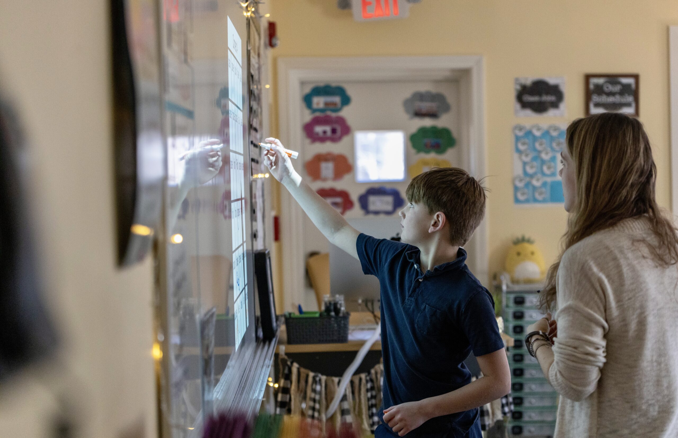 A middle school student is writing on a white board with an educator standing beside him.