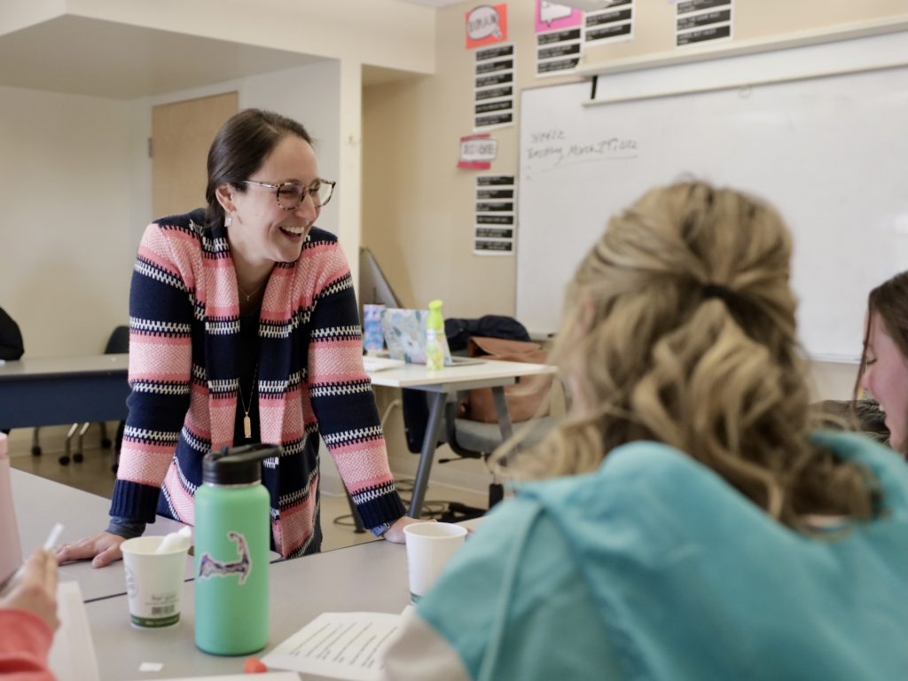 A teacher wearing glasses and a pink-and-navy striped sweater leans over a table and smiles while engaging with students in a classroom. The students, whose faces are partially turned away from the camera, are seated and appear to be working on an activity. There are papers, cups, and water bottles on the table. Behind the teacher is a whiteboard with writing and posters on the wall. The atmosphere is warm and interactive.