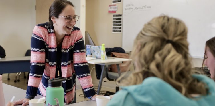 A teacher wearing glasses and a pink-and-navy striped sweater leans over a table and smiles while engaging with students in a classroom. The students, whose faces are partially turned away from the camera, are seated and appear to be working on an activity. There are papers, cups, and water bottles on the table. Behind the teacher is a whiteboard with writing and posters on the wall. The atmosphere is warm and interactive.