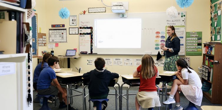 Elementary school classroom scene featuring a female teacher standing by a whiteboard, interacting with students seated at various tables. The room is brightly decorated with educational posters, a visible classroom agenda, and a projector displaying content. The students, wearing casual clothing, are attentively engaged with the teacher's presentation.