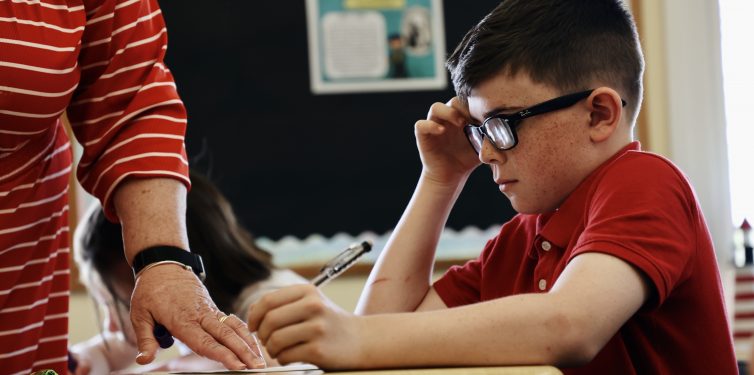 Photo of a young boy in a classroom wearing glasses, dressed in a red polo shirt, deeply focused on his work. An adult, wearing a red and white striped shirt, assists by guiding his hand as he writes. The background shows educational posters and a blackboard, indicating an engaging and supportive learning environment.