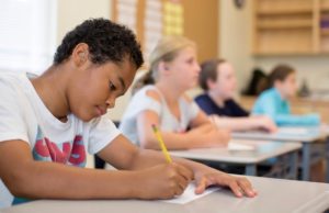A teenaged boy writes with a pencil at a desk in a classrom
