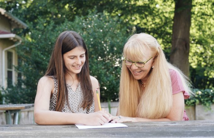 A smiling student and teacher sit side by side working on a writing task.