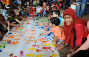 Students draw different color stick figures on a canvas banner for social awareness and unity