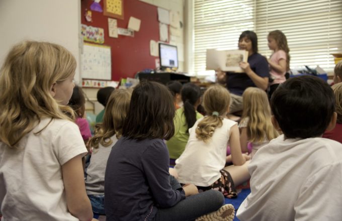 Elementary students are seated on a rug listening to an educator reading a picture book.