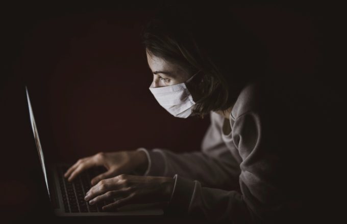 Masked student working on a laptop in a dark room.