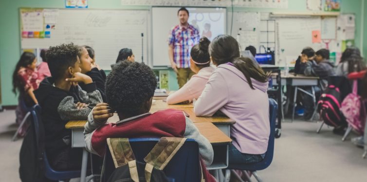 Elementary school classroom scene with diverse students attentively listening to a male teacher standing in front of a whiteboard. The classroom is decorated with educational posters and charts, and students are seated at tables in groups, contributing to a dynamic learning environment.