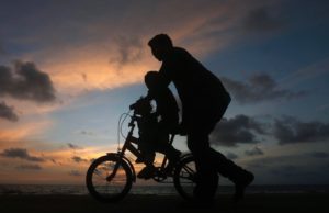 A larger and smaller child on a bicycle with a sunset behind them