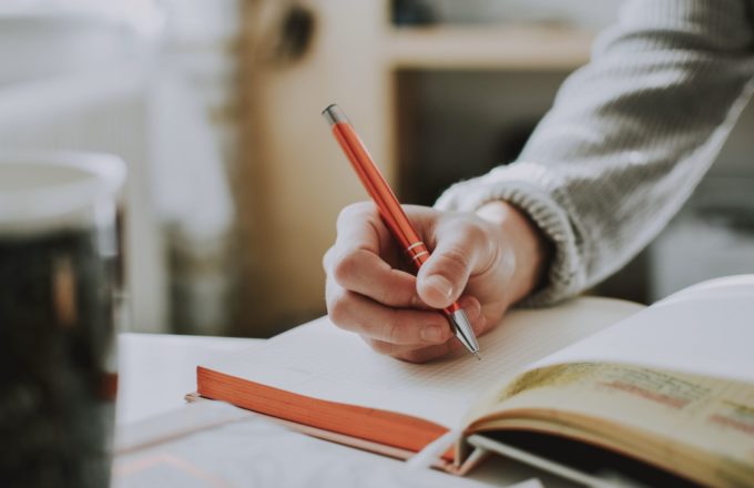 A hand holding a pen is positioned over a notebook.