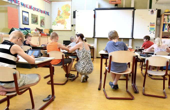 A classroom of elementary students are working independently.  An educator is kneeling next to one students' desk to provide individual support.