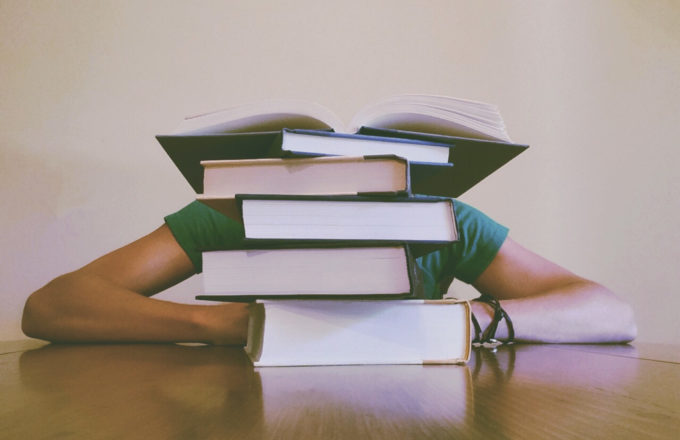 A student's face is covered by a large stack of books.